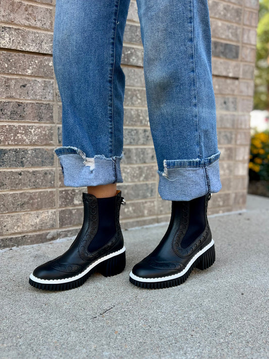Person wearing black Chelsea boots and blue jeans standing against a brick wall.