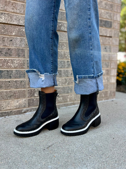 Person wearing black Chelsea boots and blue jeans standing against a brick wall.